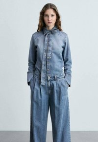 Farkkutakki, jossa on sidottu kaulus, vaalea sininen väri ja napitettava etuosa. Yhdistetty löysästi istuviin, korkeavyötäröisiin farkkusuuriin.