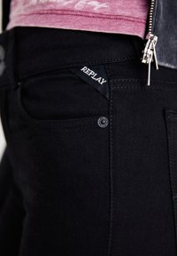 Lähikuvassa musta Replay-farkkujen tasku, jossa näkyy kangastagi ja metallinen niitti, yhdistettynä vaaleanpunaiseen yläosaan, jonka vetoketju on osittain näkyvissä.