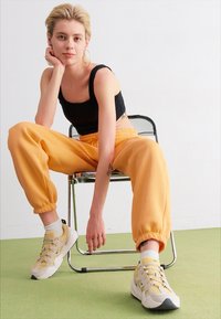 Svart avskjært tanktop, oransje tapered bukser og beige sneakers med gule aksenter. Lyse tremøbler mot en grønn gulv.