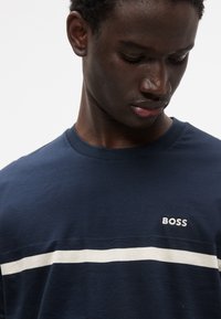 Μπλε μπλουζάκι ναυτικού τύπου με υφασμάτινη υφή, που διαθέτει μια λευκή οριζόντια ρίγα και τη κεντημένη λέξη «BOSS» στο αριστερό στήθος.