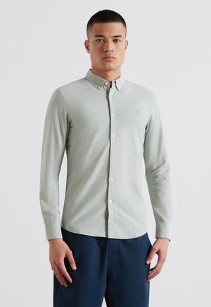 Homme portant une chemise gris clair boutonnée à manches longues et un pantalon bleu foncé, debout devant un fond blanc uni.