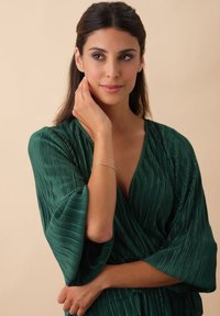 Frau mit langen dunklen Haaren trägt ein plissiertes, tiefgrünes Oberteil, berührt sanft ihr Gesicht und lächelt sanft vor einem beigen Hintergrund.