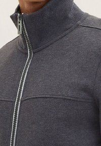 Giacca grigia con zip e colletto alto, realizzata in un tessuto testurizzato, dotata di una zip frontale con un accento metallico e cuciture piatte per un aspetto slanciato.