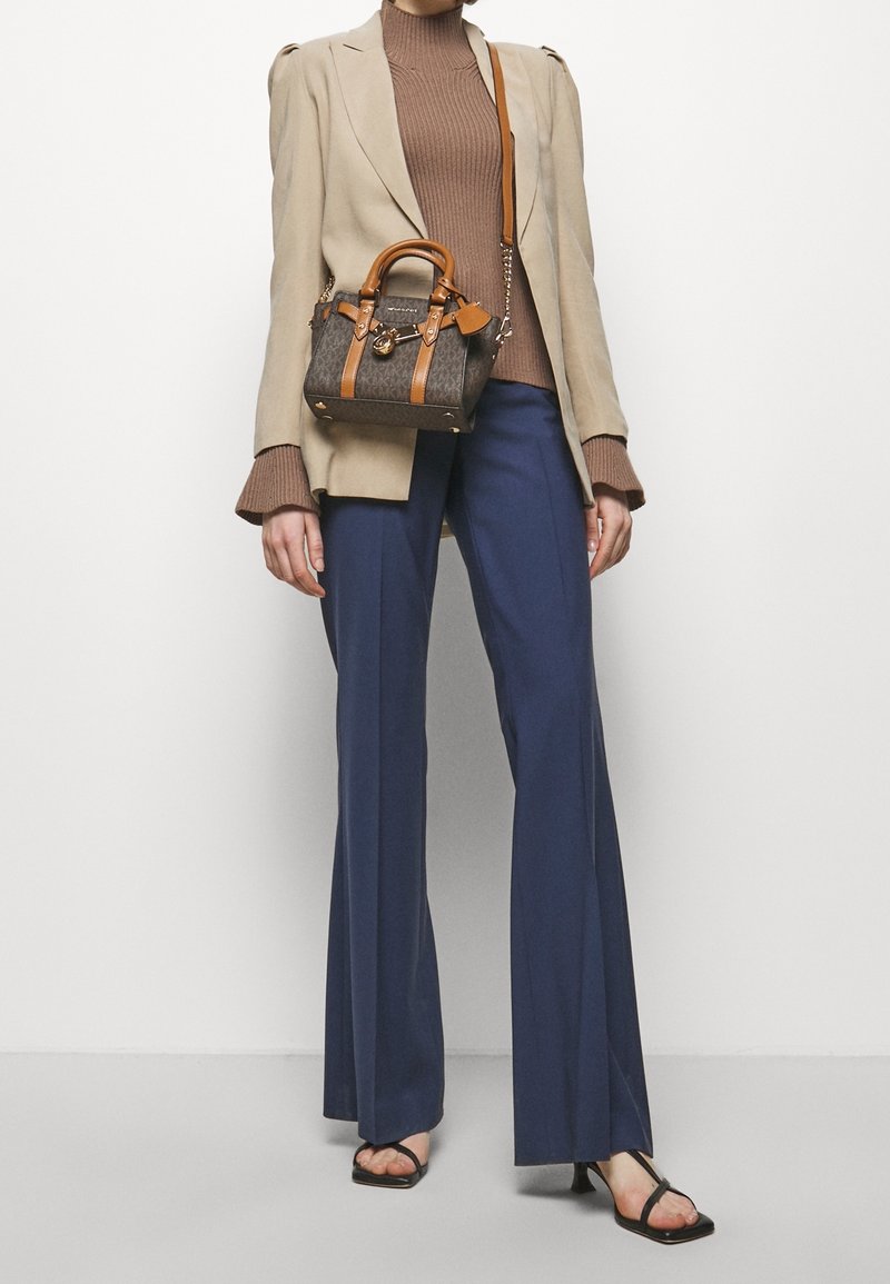Femme portant un blazer beige, un col roulé côtelé marron, un pantalon large bleu marine, des escarpins noirs à bout ouvert, et portant un sac à main marron à motifs avec des poignées couleur cuir clair.