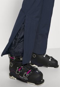 Pantaloni da sci in tessuto blu navy con cerniere ai polsini, abbinati a scarponi da sci neri con fibbie metalliche e dettagli rosa.
