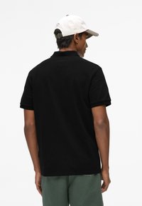 Polo noir à manches courtes, tissu texturé, avec une étiquette latérale. Porté avec un pantalon vert ; comprend également une casquette blanche et marron.