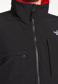 Czarna kurtka North Face z suwakiem z przodu, wysokim kołnierzem wyłożonym na czerwono oraz zapinaną na zamek kieszenią na piersi.