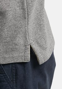 Camisa de tela texturizada gris con laterales ventilados y un dobladillo ligeramente inclinado, combinada con pantalones azul oscuro. Enfoque en los detalles del material y el corte.