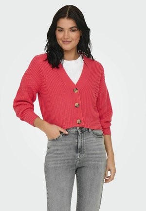 Cardigan tricoté rose corail avec un col en V, des boutons en bois et des manches ballon 3/4, associé à un jean gris taille haute. Texture douce.