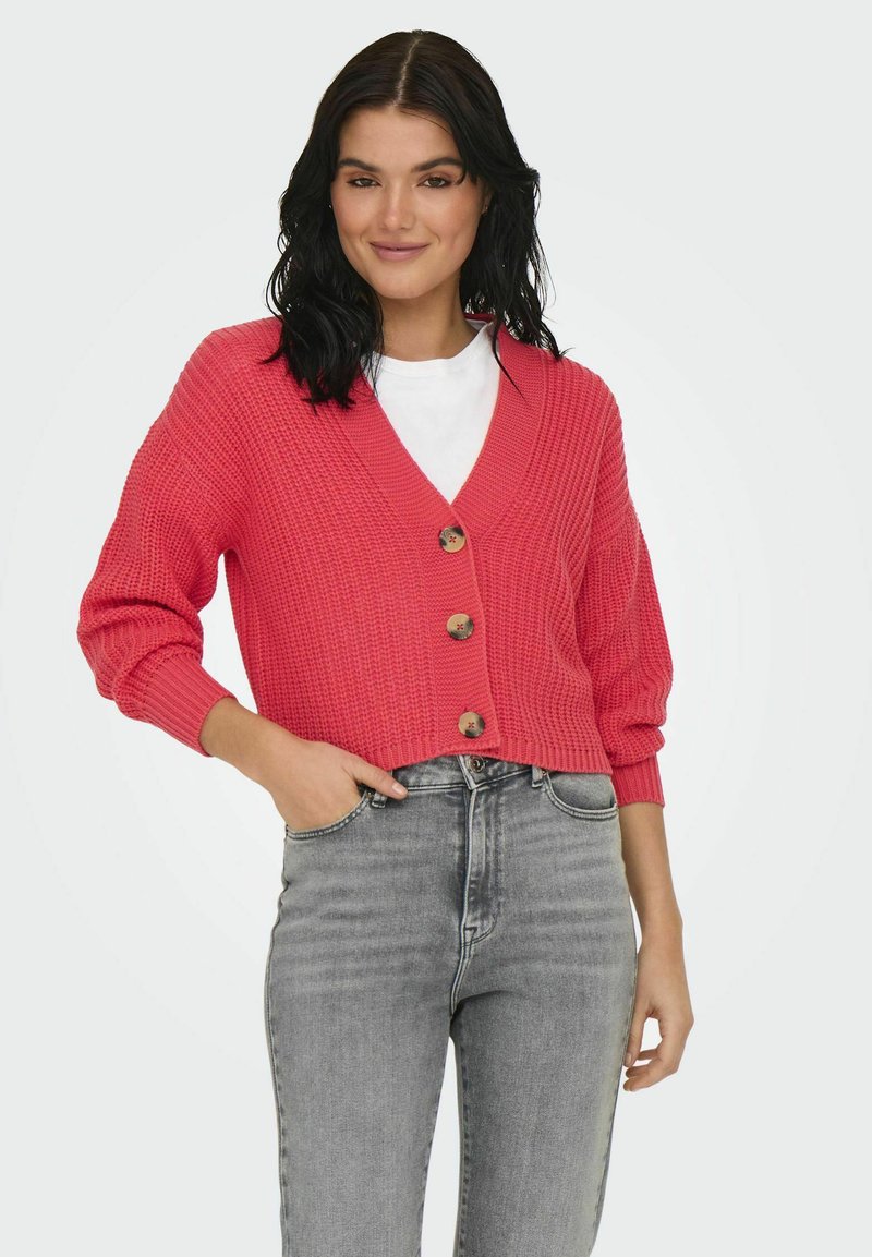 Koraalroze gebreide cardigan met een V-hals, houten knopen en driekwart ballonmouwen, gecombineerd met hoog-getailleerde grijze jeans. Zachte textuur.