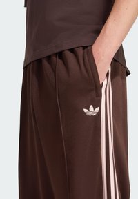 Brązowe spodnie sportowe o lu�źnym kroju, z różowymi paskami po bokach i białym logo Adidas na lewej nodze. Wykonane z miękkiego, teksturowanego materiału.