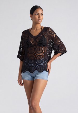 Vrouw draagt een zwarte doorschijnende gehaakte top over een zwarte bikinitop en lichtblauwe denimshorts, staand tegen een effen achtergrond.