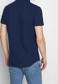 Polo blu navy a maniche corte, con colletto classico, cuciture discrete e dettagli a coste sull'orlo delle maniche. Indossata con jeans in denim chiaro.