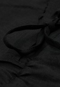 Svart tyg med en texturerad yta. Har en ögla för knytning och rynkdetaljer, som visar upp en ren, minimalistisk design.