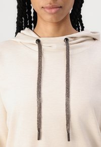 Hoodie de um tom bege claro com capuz ajustável por cordão. Os cordões são metálicos e texturizados, com extremidades metálicas escuras. Material de algodão suave.