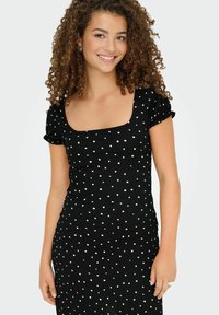 Jeune femme aux cheveux bouclés portant une robe noire à pois blancs avec de courtes manches volantes, souriant devant un fond uni.