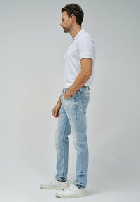 Jeans de corte slim en azul claro desgastados, combinados con una camiseta polo blanca y zapatillas blancas. Los jeans presentan desgastes y un aspecto descolorido.