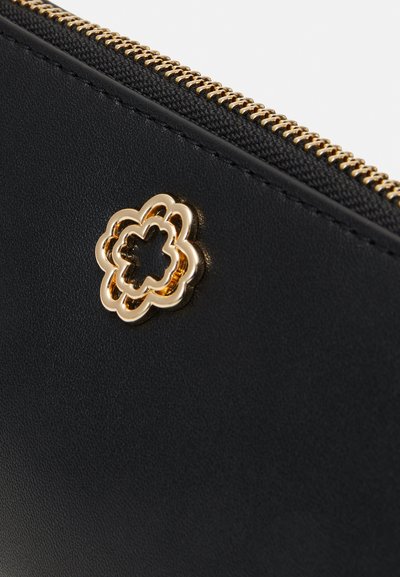 Svart skinnlommebok med et gullfarget blomsterformet emblem og gullglidelås. Teksturen er glatt, med et enkelt og elegant design.