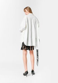 Camisa blanca de gran tamaño con textura floral, superpuesta sobre una falda negra con pliegues. Zapatos de tacón negros y un acento de etiqueta en la falda.