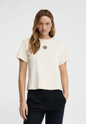 Kobieta z długimi falowanymi włosami, ubrana w kremowy krótki rękaw z małym ciemnym emblematem, czarne spodnie, stojąca z jedną ręką w kieszeni.