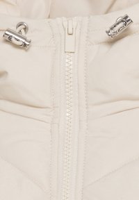 Close-up of white fabric with central zipper and two silver metal toggles attached to white cords on each side.