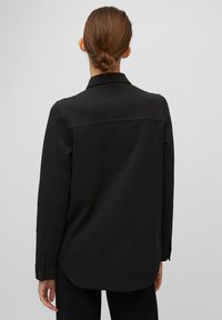 Černá košile s dlouhým rukávem a límečkem, detaily na zadním rameni a manžetami na knoflíky. Má hladkou strukturu a volný střih.