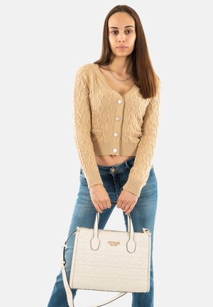 Cardigan en tricot beige avec une texture à motifs et de grands boutons, associé à un jean bleu et un sac à main matelassé blanc avec des bretelles.