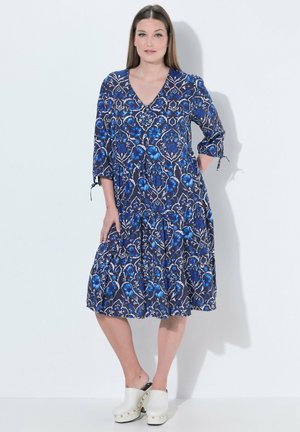 Frau trägt ein blaues, knielanges Kleid mit Blumenmuster und 3/4-Ärmeln sowie weiße Plateau-Holzclogs, steht vor einem schlichten, hellen Hintergrund.