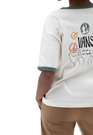 Osoba w białej koszulce Vans z zielonym wykończeniem oraz grafiką dłoni, symbolu pokoju, globu i napisem "wszystko jest w naszych rękach" na plecach.