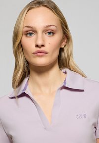 Jasnofioletowa koszulka polo z kołnierzykiem i małym haftowanym logo na piersi. Gładka faktura o luźnym kroju.