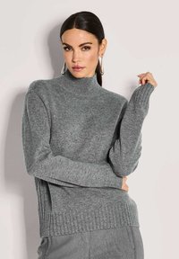 Frau mit dunklem Haar, die silberne Ohrringe und einen grauen gerippten Rollkragenpullover trägt, posiert vor einem schlichten hellen Hintergrund.