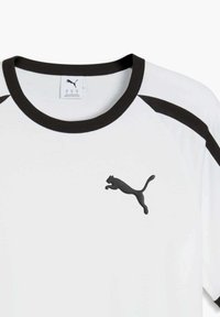 T-shirt blanc avec manches raglan noires et col noir, arborant un petit logo Puma noir sur la poitrine. Tissu doux, design minimaliste.