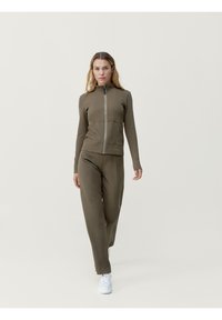 Olivovo zelená, ribovaná bunda na zips s priliehavým strihom a bočnými vreckami, doplnená o zodpovedajúce ribované nohavice a biele tenisky.