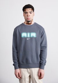 Gray sweatshirt with round neckline, featuring layered blue "AIR" text and a small Nike logo. Smooth fabric with ribbed cuffs and hem.
