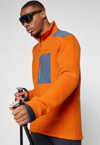 Fleece jacket in bright orange with a blue chest panel, zippered pocket, and black accents. Features high collar and textured fabric.