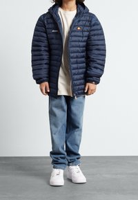 Marinblå puffjacka med horisontellt quiltat mönster, huva och logotypapplikation. Sammanvävd med ljusblå jeans och vita sneakers.