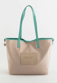 Suuri beige-kantokassi, jossa on teal-väriset hihnat, ja jossa on suorakulmainen beige-tunnus, jossa lukee "FRIENDLY PROJECT STYLE BAG". Sujuva pinta.