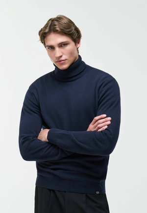 Junger Mann mit hellbraunen Haaren, der einen dunkelblauen Rollkragenpullover trägt, steht mit verschränkten Armen vor einem schlichten hellen Hintergrund.