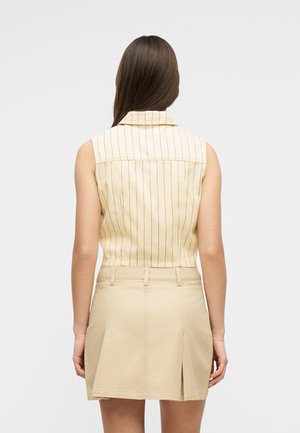 Γυναίκα με μακριά καστανά μαλλιά που φοράει μπεζ γιλέκο χωρίς μανίκια με ρίγες και ταιριαστή μπεζ φούστα, βλέποντας από πίσω.