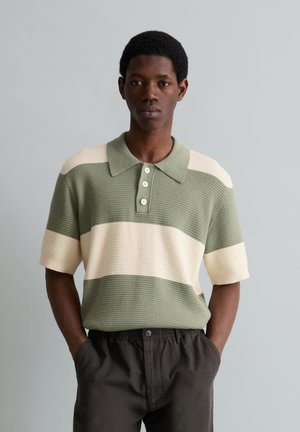 Man wearing a green and cream striped short-sleeve knit polo shirt and dark pants, standing with hands in pockets against gray background.