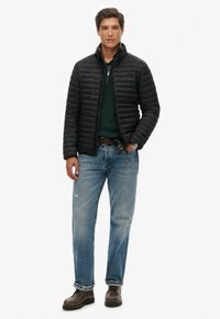 Chaqueta negra acolchada con textura acolchada, suéter verde debajo, vaqueros azul claro y zapatos marrón oscuro. El modelo posa con confianza.