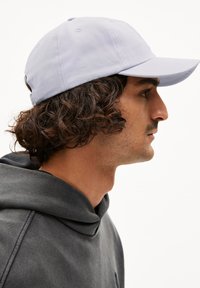 Lichtpaarse pet met gebogen klep, gemaakt van textuurstof. Het model draagt een donkergrijze hoodie, haar zichtbaar onder de pet.