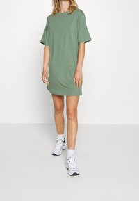 Zielona bawełniana sukienka t-shirtowa o luźnym kroju, z krótkimi rękawami i okrągłym dekoltem. Zestawiona z białymi sportowymi butami i skarpetkami.
