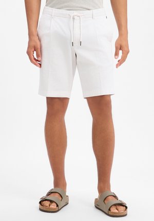 Mann trägt weiße knielange Shorts mit Kordelzug in der Taille und graue Sandalen mit Schnalle, steht vor weißem Hintergrund.