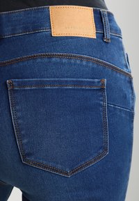 Modré džínové kalhoty s hladkou texturou, s hnědou koženou nášivkou označenou "VERO MODA" a viditelnými švy.
