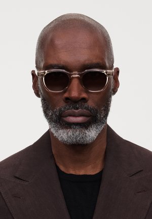 Skallet mann med grå skjegg som har på seg gjennomsiktige runde solbriller og en mørkebrun blazer over en svart skjorte, nøytralt uttrykk.