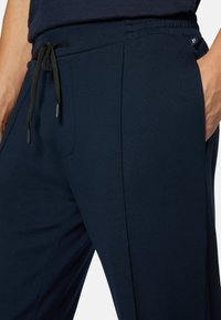 Man klädd i marinblå joggerbyxor med svart dragsko, händerna i fickorna, närbild på midja och övre ben.