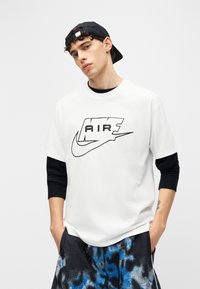 Νέος άνδρας που φοράει λευκό μπλουζάκι Nike Air πάνω από μαύρο μακρυμάνικο πουκάμισο, μαύρο καπέλο με την ανάποδη φορά, και μπλε-μαύρο σορτς με σχέδιο, με τα χέρια στις τσέπες.