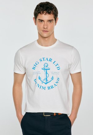 Mężczyzna w białej koszulce z niebieską grafiką kotwicy i napisem "Big Star Ltd Denim Brand" oraz ciemnych spodniach z plecionym paskiem.