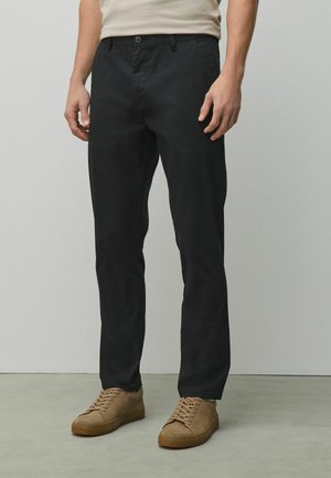 Mężczyzna ubrany w czarne proste spodnie i jasnobrązowe zamszowe trampki na sznurówki, stojący na tle gładkiej jasnoszarej ściany i podłogi.